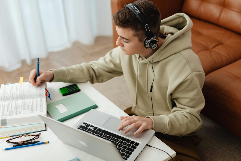 Teen studying at home using laptop and books, wearing headphones, sitting in a cozy room.